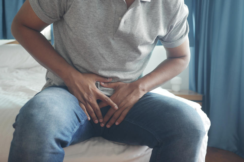 A person wearing jeans and a gray polo shirt sits on a bed holding their lower abdomen, appearing to be in discomfort. No names provided